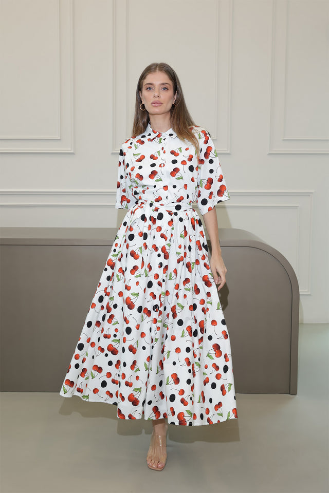 A woman standing in a room wearing a white dress with a red cherry polka dot print.